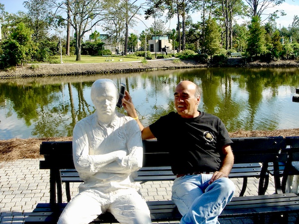 Steve sitting on a park bench next to a white sculpture of a many with arms crossed. Steve's putting his phone to the sculpture's ear.