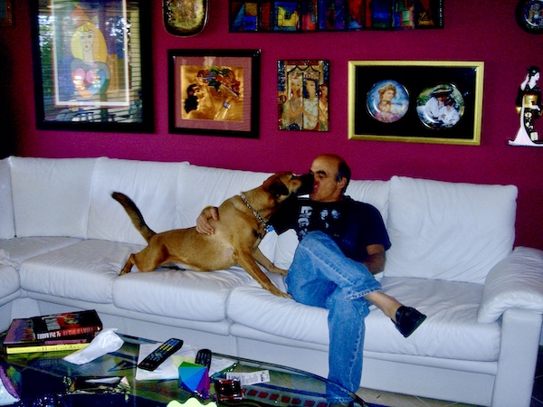 Steve on a white sofa with a tan dog licking him.