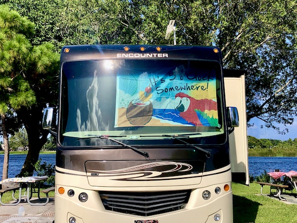 A brown and tan RV with a colorful sign in the window that says It's 5 o'clock somewhere". Seen at John Prince Park Campground Lake Worth FL