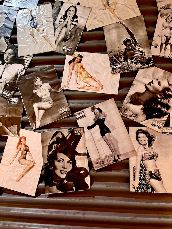 Pages of pinup girls taped to a wall at the Torn roof exposed to the sky with WW2 planes seen flying overhead