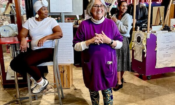 Silver haired woman in purple top with purple, green and gold leggings speaking to an audience. The woman is Liz Williams Founder SoFab