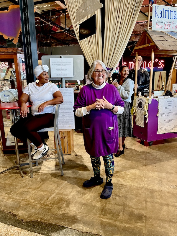 Silver haired woman in purple top with purple, green and gold leggings speaking to an audience. The woman is Liz Williams Founder SoFab
