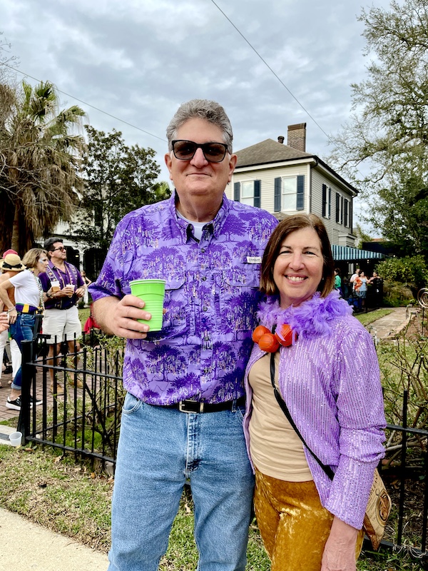 Tall man with sunglasses and a purple shirt with a woman in a lavender jacket with purple feather boa at the 52 Year Mardi Gras Tradition