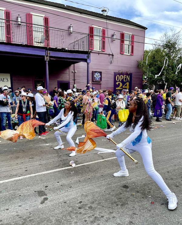 At Our First Mardi Gras Parade girls in white costumes with orange flags dance in the street.