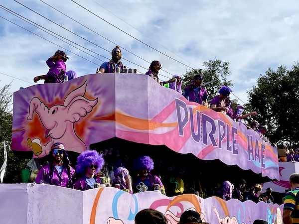 Pink and purple float with purple haired people through Mardi Gras beads.