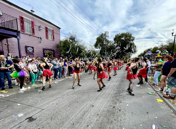 Large group of women in black tops and short red skirts dancing down the street at Our First Mardi Gras Parade