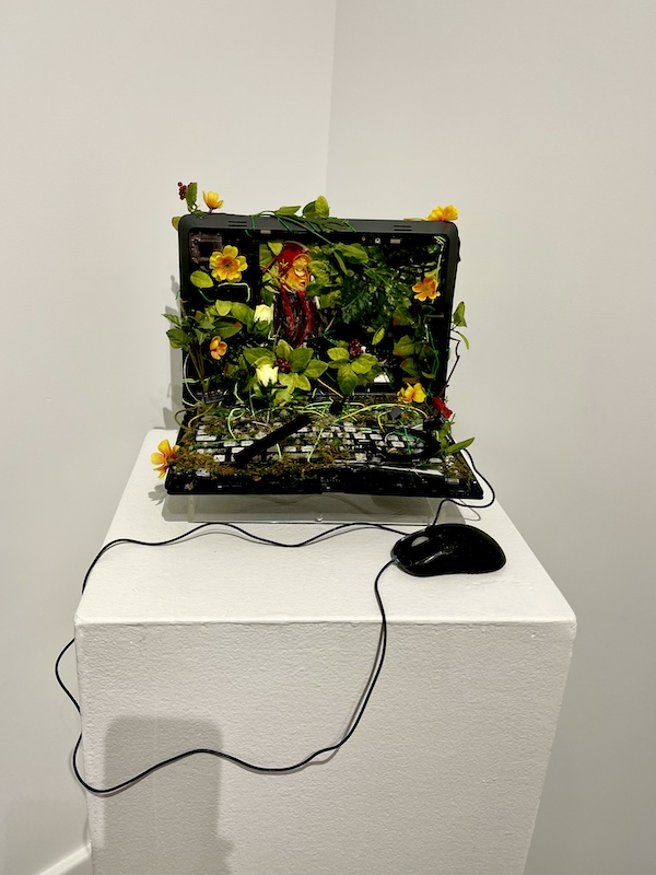 Black laptop computer with silk flowers and leaves. Attached is a mouse. On display at The Grace Museum Abilene TX