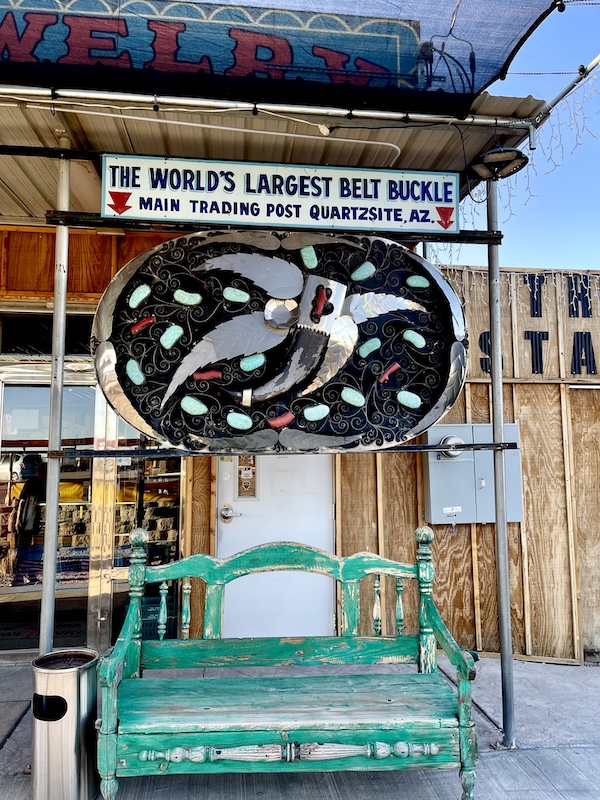 Huge black, silver, turquoise and coral belt buckle hanging above a green bench seen on Our Visit to Quartzite Arizona