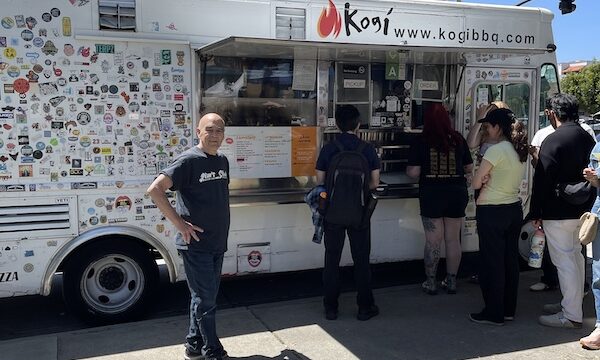 White food truck with colorful stickers and Steve getting in line to order food.
