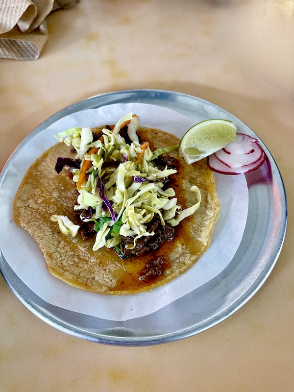 Corn tortilla topped with steak, sauce, and slaw with lime wedge and radish slices at Trejo's Tacos Hecho en Los Angeles