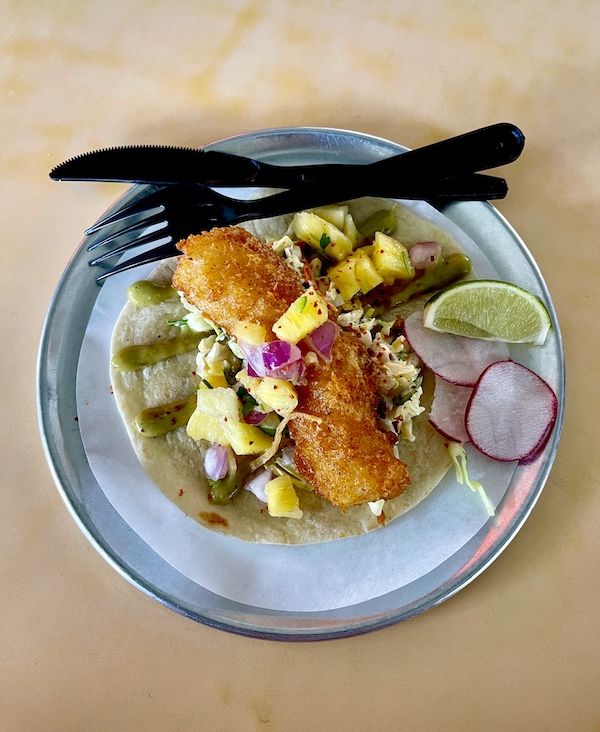 Tortilla topped with slaw, crispy battered fish, avocado sauce and pineapple chunks with lime wedge and radish slices at Trejo's Tacos Hecho en Los Angeles