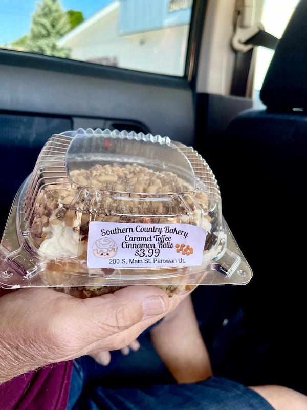 Man's hand holding a closed plastic clam shell filled with an iced caramel toffee cinnamon roll bought in Delightful Parowań Utah