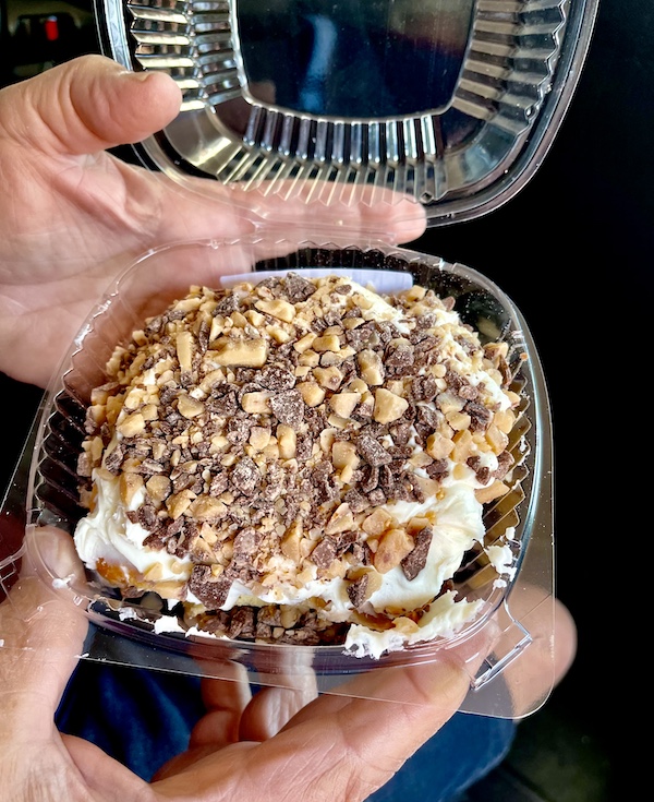 Man's hand holding an open plastic clam shell filled with an iced caramel toffee cinnamon roll bought in Delightful Parowań Utah