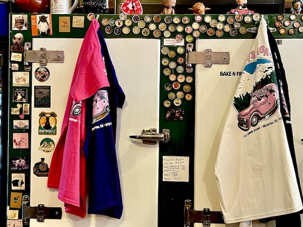 T-shirts and aprons on display from 2 antique refrigerators at Thee Pitts Again BBQ. Lots of bottle cap magnates around the fridge doors.