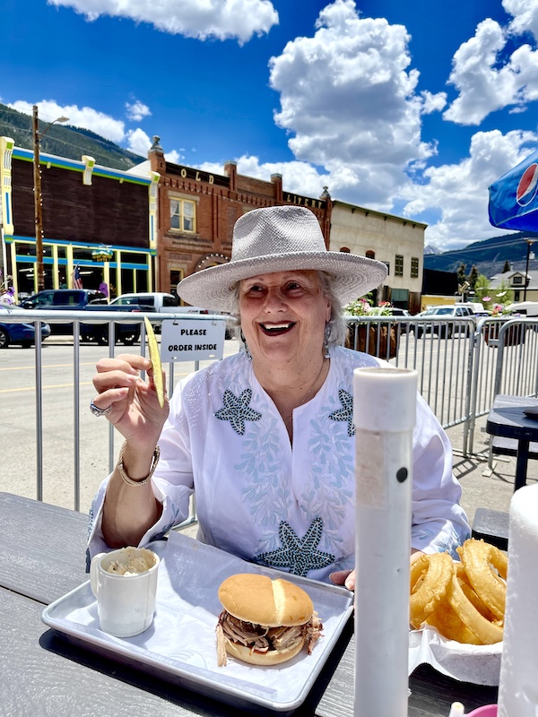 Woman in a gray hat holding a thin pickle spear eating outdoors at Thee Pitts Again BBQ