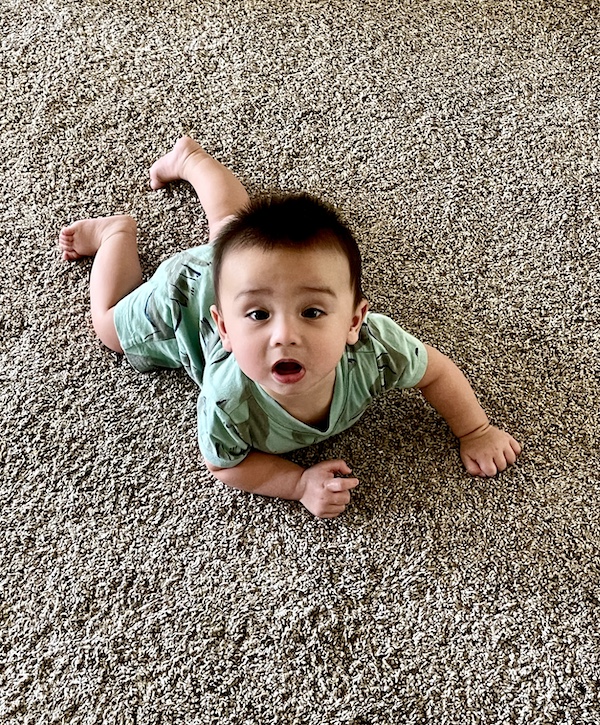 Baby crawling on carpet in a green romper on a beige carpet.