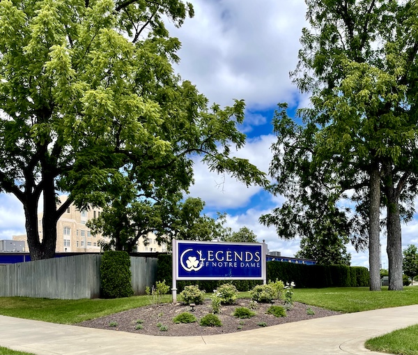 Blue sign on lawn between trees with building in background seen during our fun time at Notre Dame