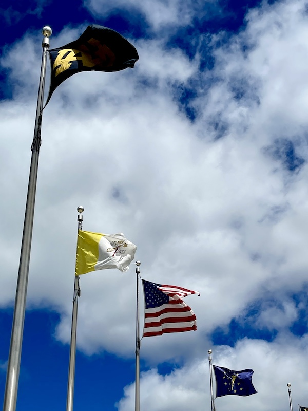 A series of flags flying above Notre Dame's football stadium with lots of cloud and deep blue sky.
