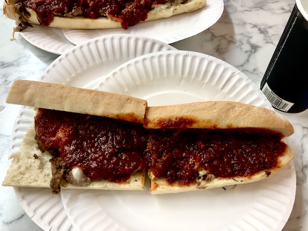 East Coast Here We Come. Cheesesteak with sauce sitting on white paper plates and cut in half.
