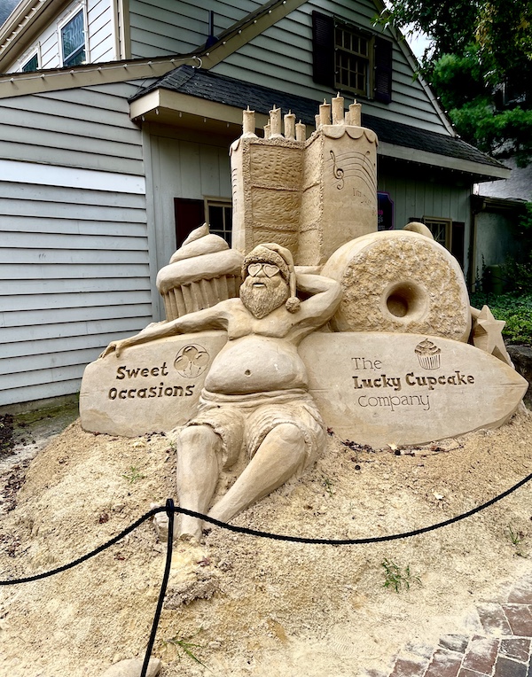 The last of the Sand Sculptures Peddler's Village depicts Santa , topless in shorts with sunglasses and a Santa hat sunbathing in front of a towering cake, cupcake and donut.