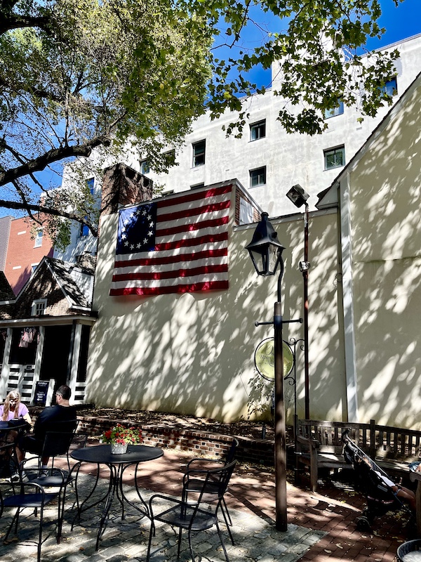Courtyard at Betsy Ross' house that has a replica of the original Stars and Stripes hanging on a white building wall. Metal tables and chairs are scattered in the yard.