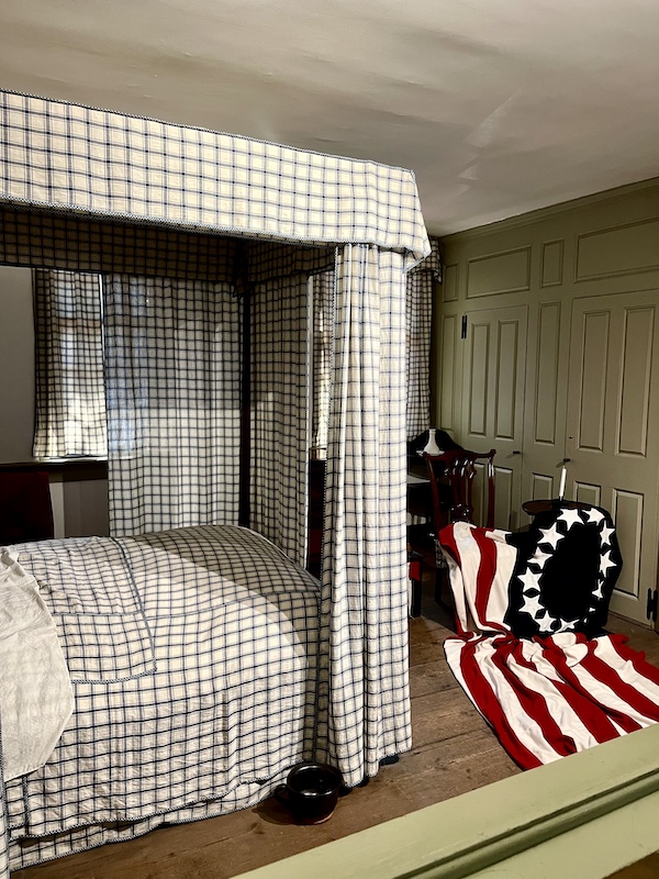 Canopy bed with white and black grid pattern drapery, American flag on floor by chair seen while Visiting Betsy Ross' House