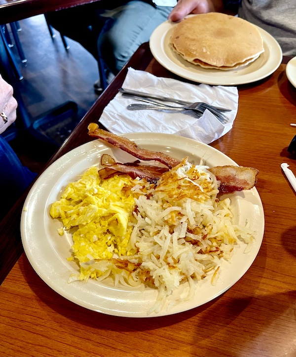 Beige plate topped with scrambled eggs bacon and potatoes and one topped with pancakes on a wood tabletop at Jimmy's Family Restaurant Easley SC