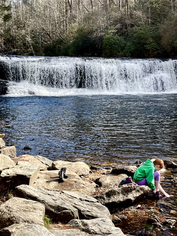Red haired little girl sitting on a rock by Hooker Falls, with her bare feet in the water. Her cowgirl boots are on another rock.