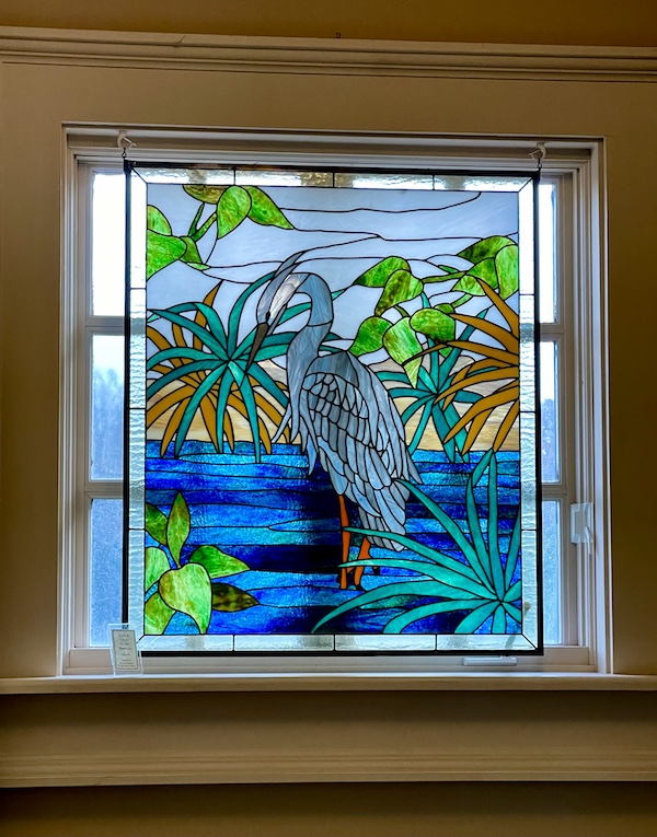 Stained glass window in blues, grays and greens of a bird in water with tropical greenery. Seen on our South Carolina College Tour