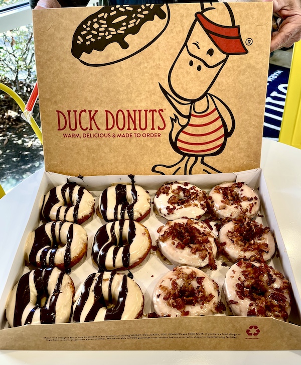 Box of 12 donuts with 6 covered in tan glaze and dark brown drizzle. The other 6 are maple glazed with lots of bacon pieces.