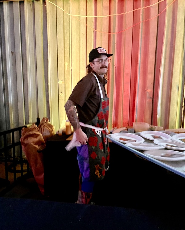 Man with mustache and black cap standing behind a table with plates of food at Family Style FoodFest NOLA