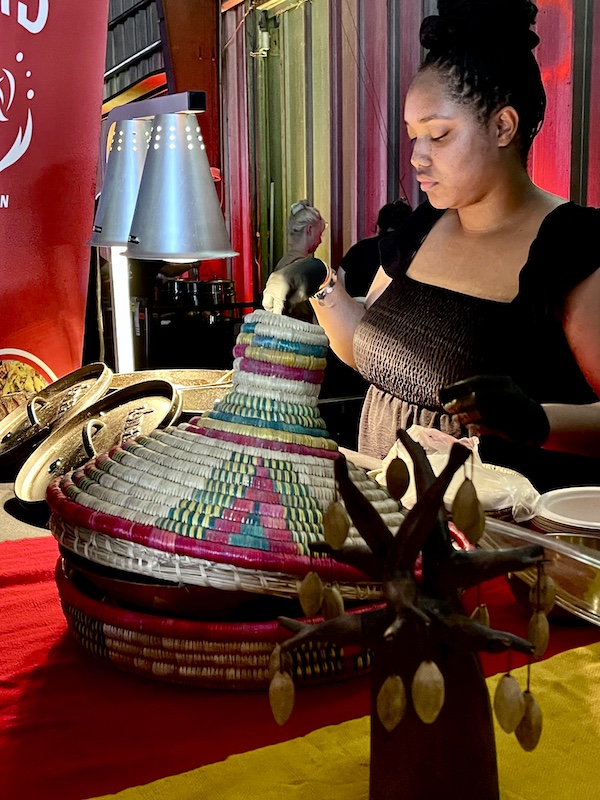 Woman serving food and a display of African baskets at Family Style FoodFest NOLA