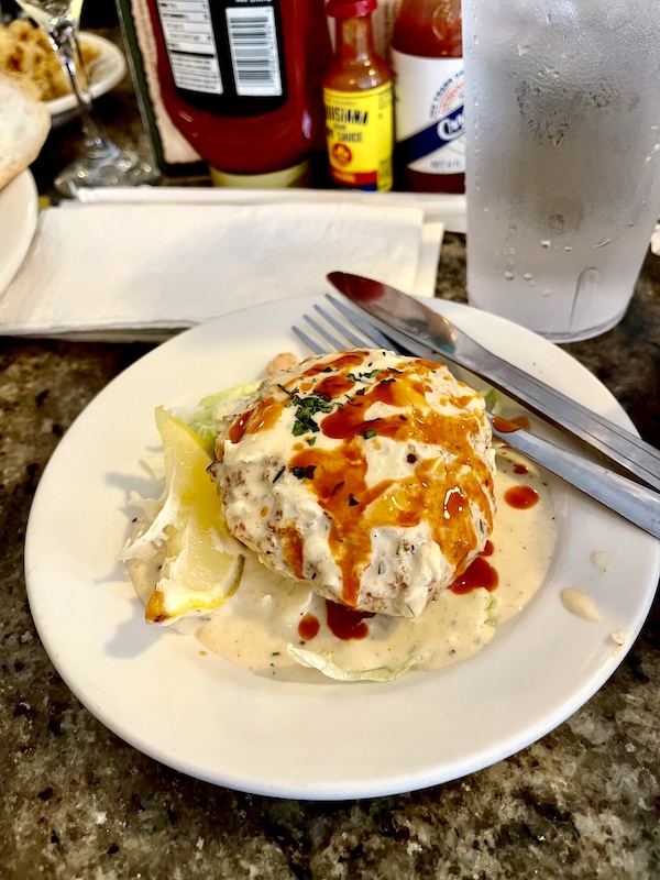 Crab cake covered in white sauce and a drizzle of red hot sauce on white plate with knife askew. My entree for our French Quarter Girls Lunch