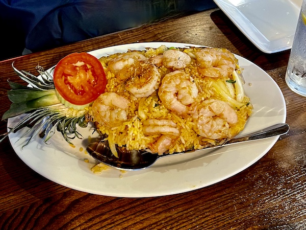 Half a hollowed pineapple filled with fried rice, veggies, pineapple chunks and shrimp with a tomato slice garnish on a white oval plate at Thaicoon Restaurant Pearland Texas