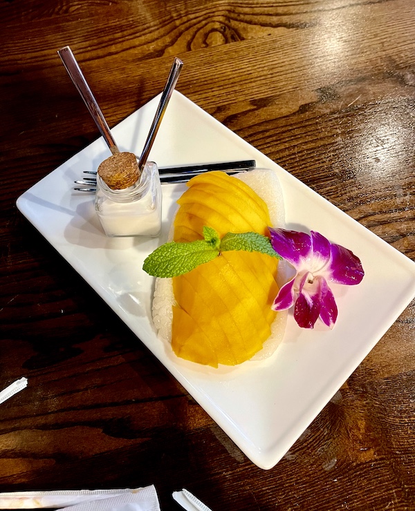 Rectangular white plate topped with rice, mango slices, jar of coconut milk and a purple orchid flower at Thaicoon Restaurant Pearland Texas