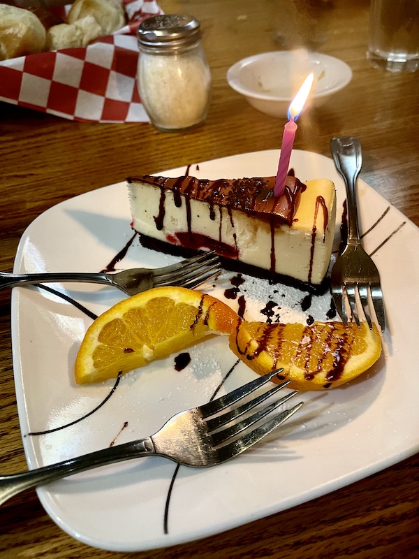 Plate topped with wedge of cheesecake drizzled with chocolate sauce and a birthday candle. Also on the plate at Italian Garden Restaurant Canyon Lake TX is a twisted orange slice and fork.