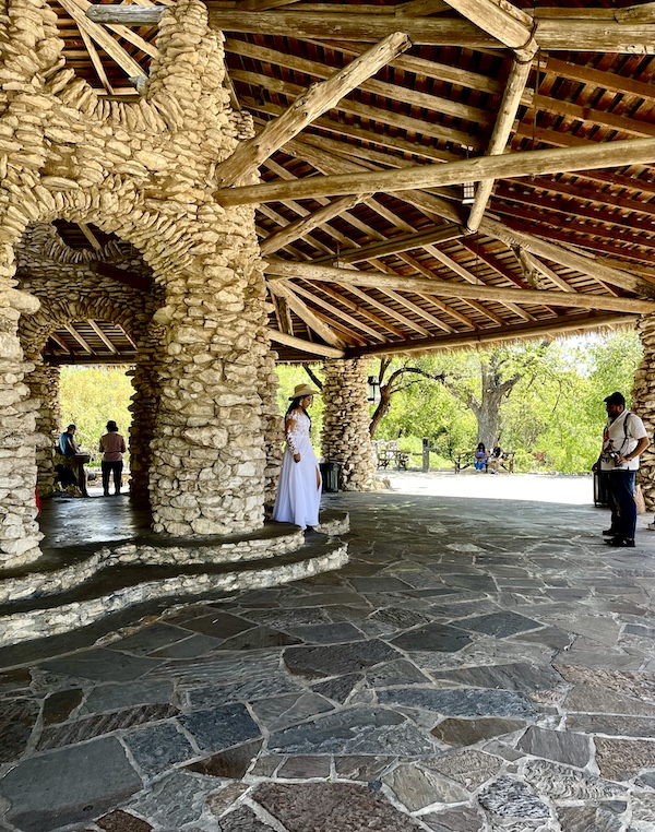 Photographer taking pictures of a girl in white gown and cowboy hat under limestone and wood pagoda at Japanese Tea Garden San Antonio
