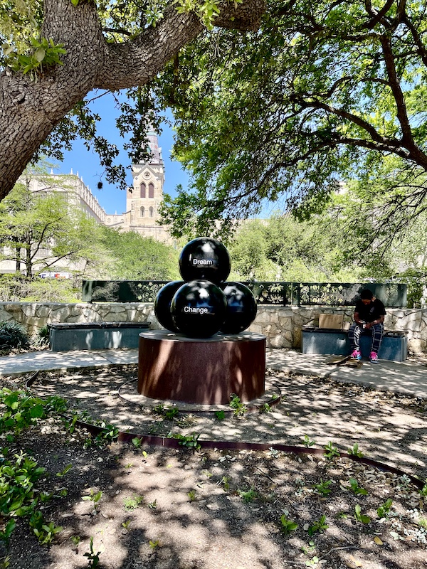 Black sculpture of stacked balls with words in park by San Antonio River Walk