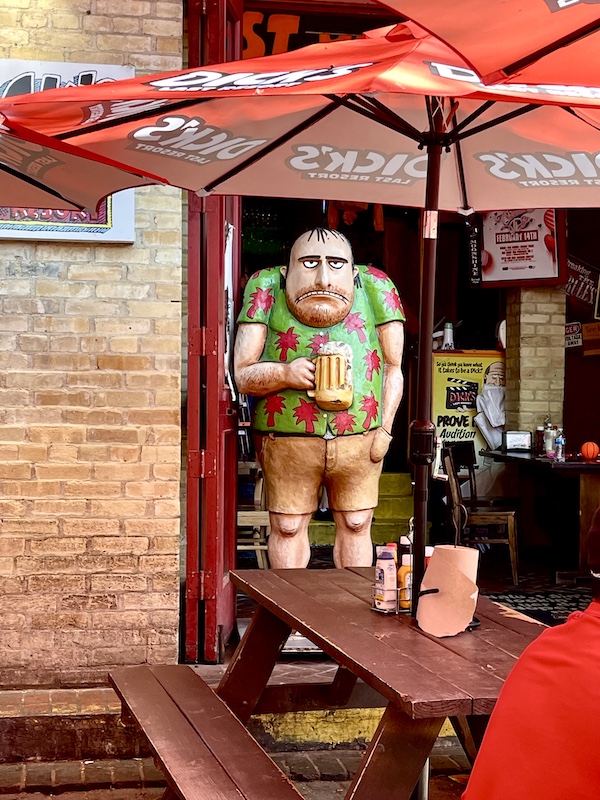 Red market umbrella and funky wood statue of a man holding a beer mug at San Antonio River Walk