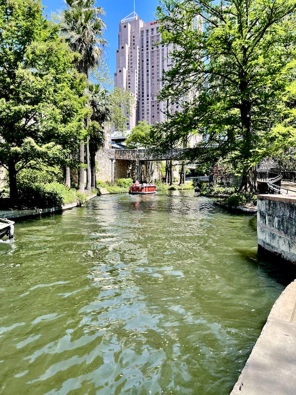 Red boat, river and tall white building seen as we stroll along San Antonio River Walk
