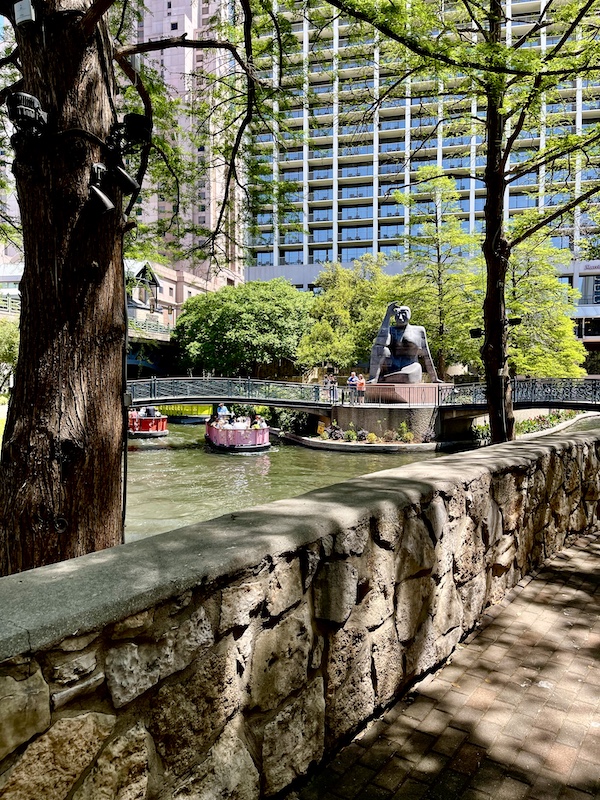 Huge statue of a woman by a bridge that spans the San Antonio River. Colorful boats cruise the river and large buildings and trees are visible.