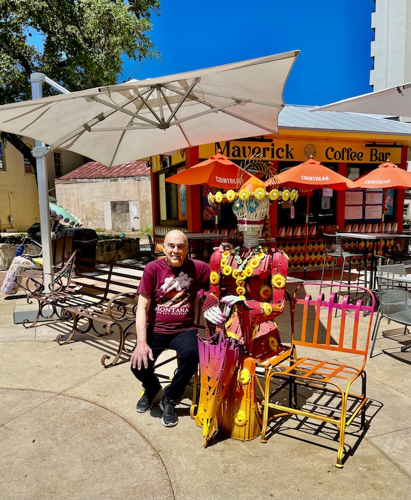 Steve on a bench sitting next to a skeleton dressed in a colorful outfit with a sombrero after our visit to San Antonio River Walk