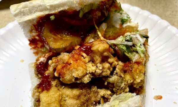 White plate topped with half of a fried oyster po' boy with hot sauce, pickles, remoulade, and shredded lettuce eaten during our Bonus Visit to NOLA