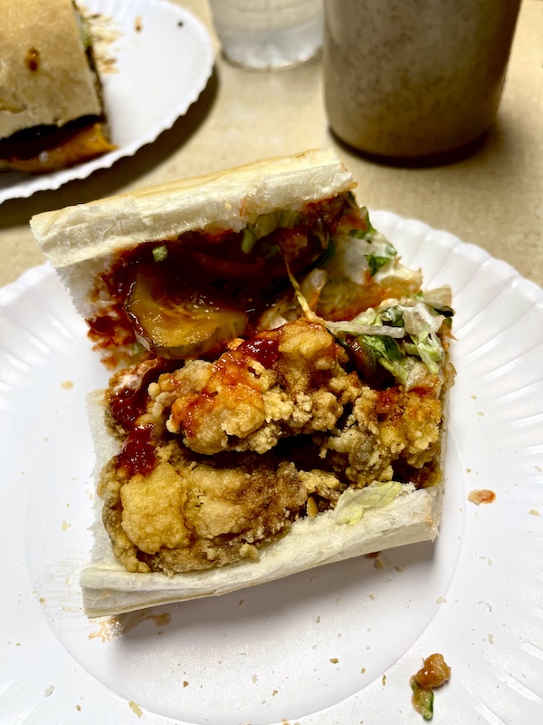 White plate topped with half of a fried oyster po' boy with hot sauce, pickles, remoulade, and shredded lettuce eaten during our Bonus Visit to NOLA