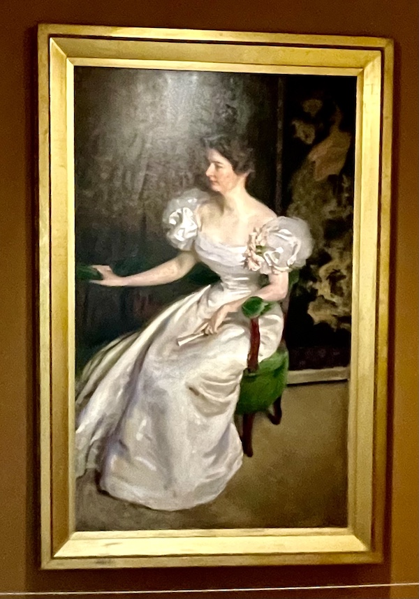 Painting of a woman in a blush satin gown with tuille sleeves seated in a green velvet companion chair, holding a fan at the Westmoreland Museum of American Art