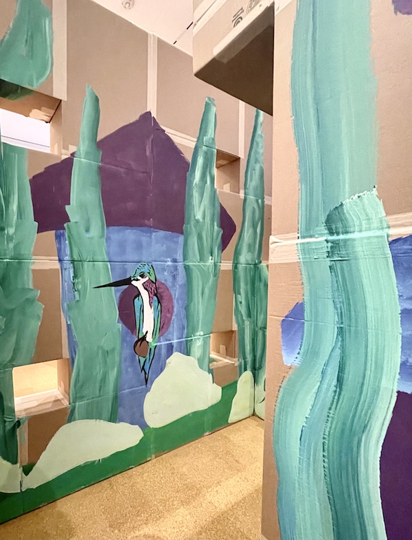 Mural of colorful bird in front of a house with stylized green trees painted on cardboard at Westmoreland Museum of American Art 2