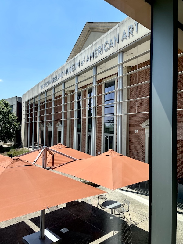 View of the front of the Westmoreland Museum of American Art and several orange market umbrellas though a glass window.