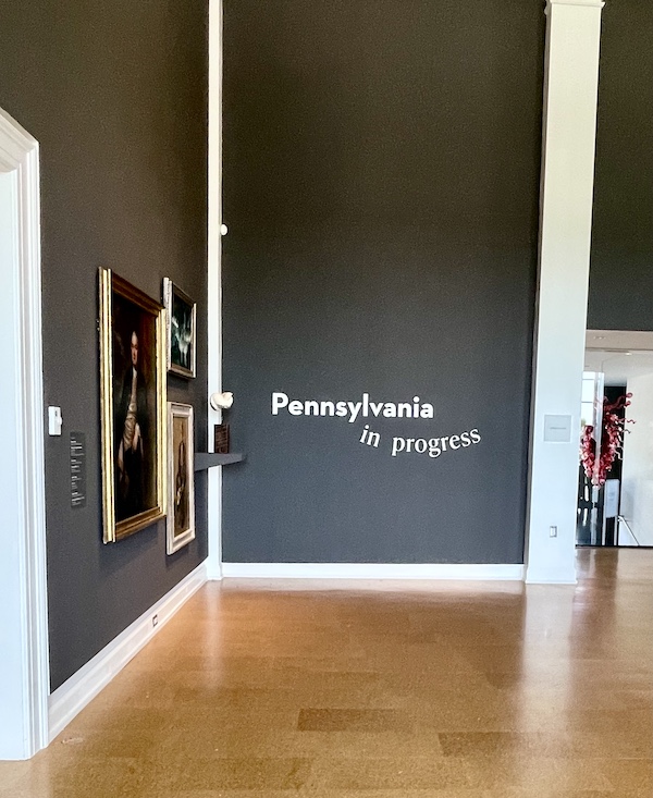 Dark gray walls with art hanging and one wall with title of exhibit at the Westmoreland Museum of American Art