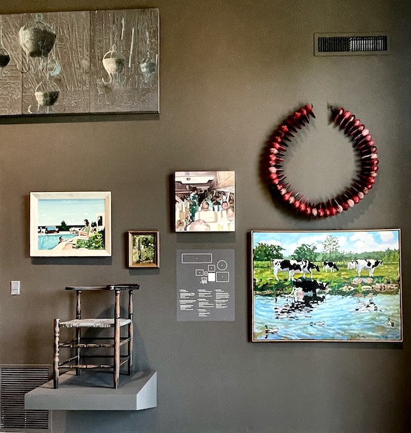 An assortment of paintings and objects displayed on a dark gray wall at the Westmoreland Museum of American Art