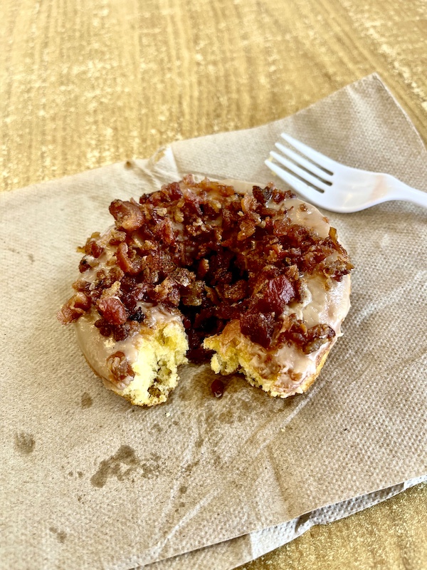 Bacon topped and maple glazed donut that has a bite missing, sitting on a napkin with a fork on a light wood table.